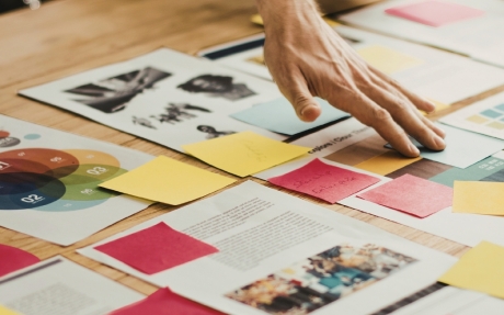 Mehrere Hände ordnen bunte Papierstücke und Farbfächer auf einem Tisch mit gedruckten Dokumenten an