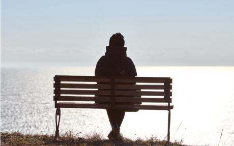 Person mit Kapuzenjacke sitzt allein auf einer Parkbank mit Blick auf das Wasser