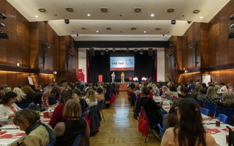 Großer Saal mit Publikum an Tischen und Bühne mit drei Personen vor Leinwand mit Schrift 'VHS-TAG'